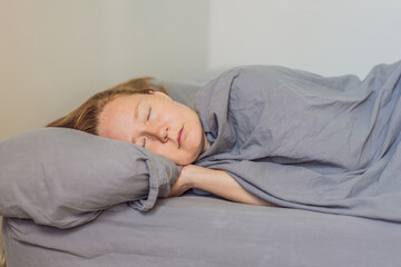 Woman peacefully sleeping in bed with soft gray bedding, enjoying a calm and comfortable morning atmosphere. Rest, relaxation and wellbeing concept, representing healthy sleep, serenity and cozy home