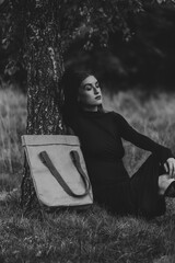 Elegant Black and White Portrait of a Woman Resting at a Tree Trunch with a Large Ecological Tote Bag Next to Her on the Grass