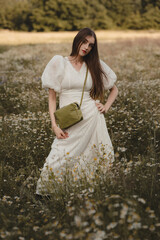 Charming Woman in Long Boho Lace Dress with Small Green Crossbody Handbag Standing Among Blooming Chamomile Meadow