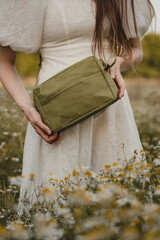 Close-up of Hands Holding a Small Green Crossbody Bag made of Ecological Washpapa Material Among Blurred Meadow Flowers in Summer