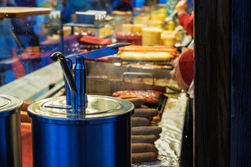 Two stainless steel condiment pumps for ketchup and mustard beside a grill filled with sausages at a christmas market food stall