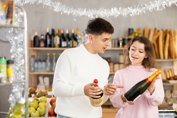 Cheerful young girl and guy in twenties excitedly discussing while comparing bottles of wine at Christmas-decorated grocery store, preparing for holiday celebration