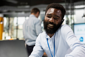 Black male employee in corporate office having phone call with coworker and typing on laptop. African american professional sharing business ideas during mobile conversation with associates.