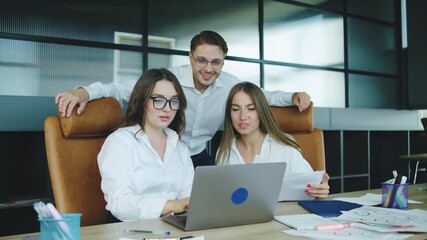 Three colleagues gather around a laptop in a modern office, surrounded by papers, cheering and high-fiving as their business plan shows positive results