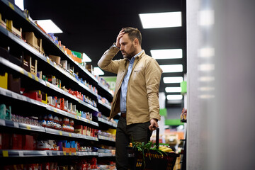 Worried man feeling shocked by grocery prices while shopping at supermarket.