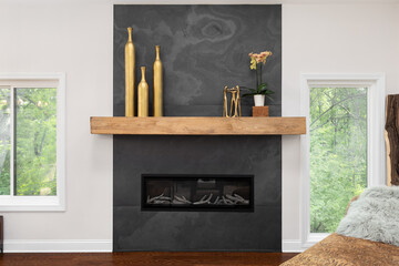 Obraz premium A modern fireplace with a dark slate surround and a natural wood mantel. Golden decorative vases and a plant add elegance with windows on the side.