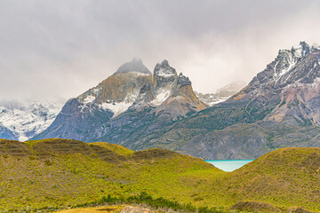 Fototapeta premium Dramatic mountain peaks rising above rolling terrain with a glimpse of turquoise water in Torres del Paine National Park, Patagonia, Chile.
