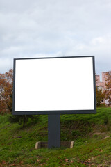 Fototapeta premium Large blank outdoor billboard standing in a green urban area, photographed on an overcast day. The empty white advertising panel is mounted on a dark metal structure, surrounded by grass, trees