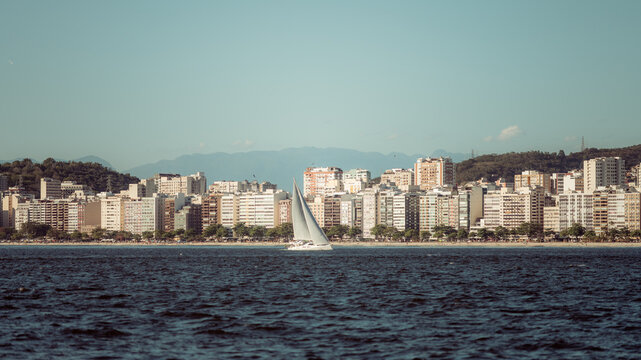 Telephoto view of Niteroi coastline with dense beachfront skyline, calm sea and a white sailboat gliding in foreground against distant soft-blue mountain silhouettes - Powered by Adobe