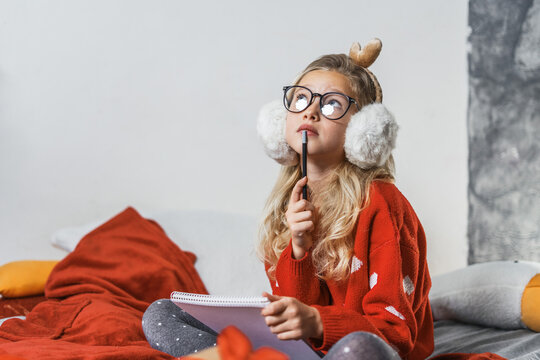 Teenage girl sist on bed, holding pencil, reflecting joyful christmas holiday inspiration. Cozy winter ambience, glitter ornaments, festive spirit help shape fresh creative ideas and future plans.
