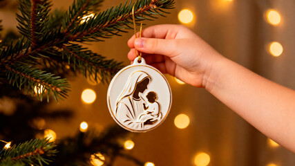 Christmas ball. Child's hand gently holds a wooden ornament depicting a mother and child, hanging on a Christmas tree adorned with soft glowing lights, celebrating the holiday spirit