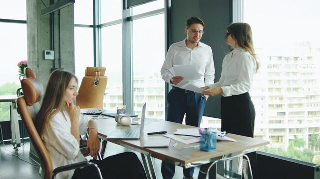 In a modern office, a man shows documents to a female colleague in frustration, while another woman continues working on her laptop at the desk