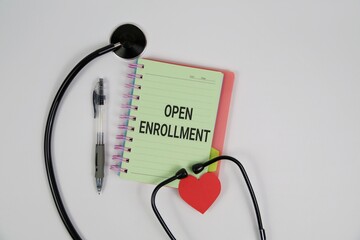 A medical-themed open enrollment concept featuring a notebook labeled 'Open Enrollment', a stethoscope, pen, and a red heart on a white backdrop, symbolizing health care choices.