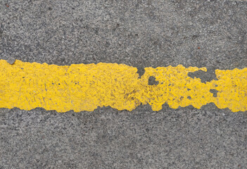 Extreme texture detail of gray asphalt pavement crossed by a vibrant yellow road line exhibiting heavy peeling and traffic wear. Excellent textured design element.