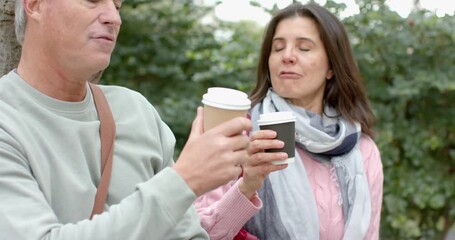 Diverse senior couple raising coffee cups on park walkway, toasting brew while chatting and sipping - Powered by Adobe