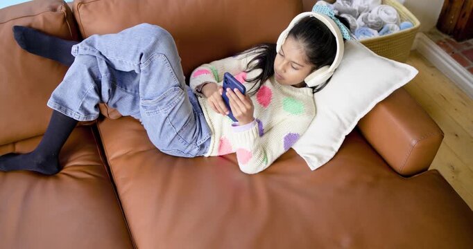 Indian girl child tapping and scrolling smartphone with headphones on couch laughing, copy space