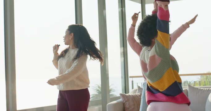 Diverse female friends dancing miming lyrics using pen mic in sunlit living room with ocean view