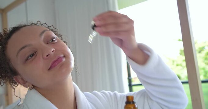 African American woman in bathrobe reaching amber dropper at vanity, applying serum for skincare