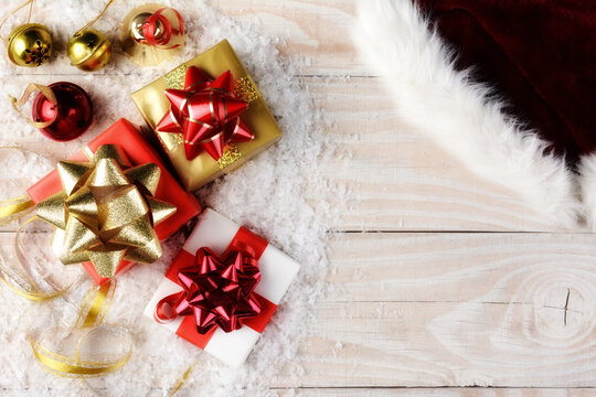 A Santa Hat Christmas presents, bells and ribbon with artificial snow on a rustic wooden table. - Powered by Adobe