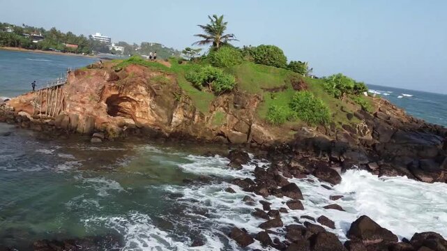 Ocean waves crashing on the parrot rock, a small island connected by a bridge to the main beach in Mirissa, with cheerful tourists walking, Sri Lanka