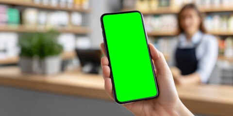 Hand holding smartphone with green screen display. Close-up of a hand holding smartphone with blank green screen in a shop or cafe environment, ready for app or design mockup.