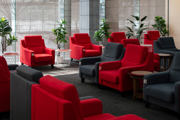 Modern lounge seating area with numbered red and dark gray armchairs, side tables, and indoor plants, illuminated by natural light through large glass windows