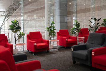 Modern lounge seating area with numbered red and dark gray armchairs, side tables, and indoor plants, illuminated by natural light through large glass windows