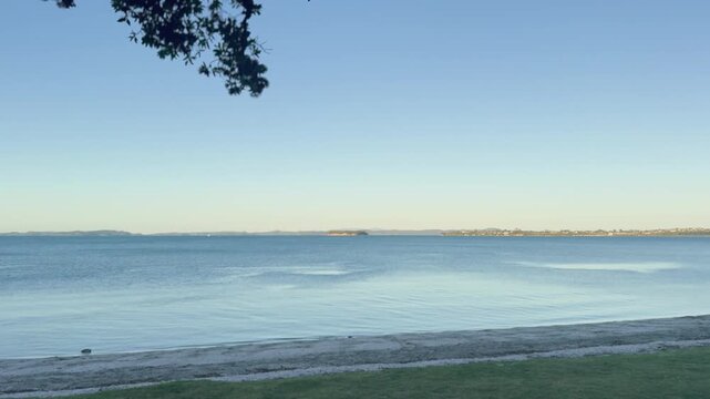 A tranquil New Zealand coastline at sunset, featuring calm ocean waters under a soft blue and orange sky. The minimal, quiet atmosphere highlights the simplicity and beauty of nature, with gentle ligh