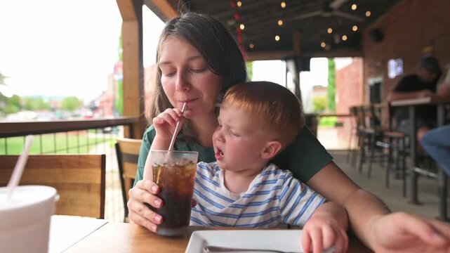 caucasian mother and toddler sipping cola through straw at outdoor table, child watches mother taste, ice clinking in glass, porch lights and casual cafe backdrop create playful, joyful