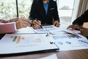 A group of professionals reviews charts on paper and a tablet, discussing financial data and calculating figures during a business meeting.