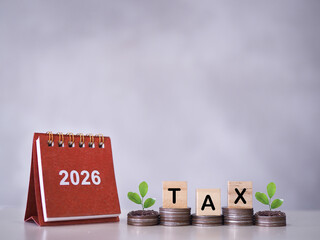 2026 desk calendar, Wooden blocks with the word TAX and plants growing up on stack of coins. The concept about saving money and manage time to paying tax in year 2026