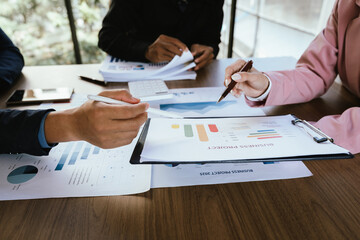 A group of professionals reviews charts on paper and a tablet, discussing financial data and calculating figures during a business meeting.