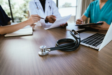 A stethoscope lies on a wooden desk while medical professionals work on a laptop in the background during a healthcare meeting.