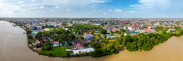 Wide aerial panorama capturing the contrast between the historic buildings of Paramaribo and the lush green vegetation along the Suriname River banks