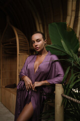 A woman in a purple robe poses near a bamboo doorway with soft natural light and tropical plants around
