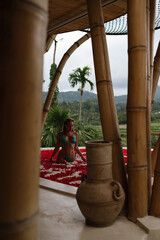 A woman sits in a flower bath surrounded by bamboo structures and tropical greenery, with rice fields visible in the background