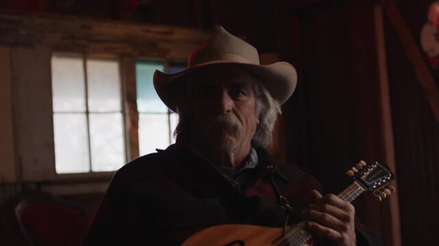 Man With Mustache Wearing Cowboy Hat Holding Mandolin String Instrument Inside of Shadowy Saloon