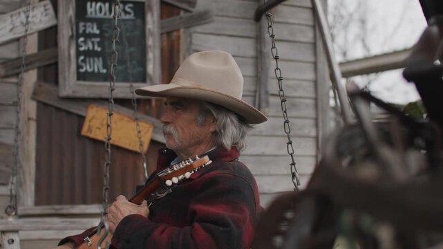 Man Wearing Cowboy Hat and Flannel Shirt Holding Mandolin String Instrument and Sitting on Porch Swing Outside of Old Rustic Saloon