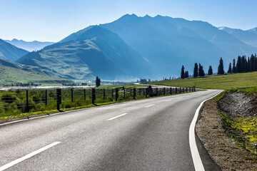 Empty asphalt highway road curving through green grassland with mountain range background under blue sky