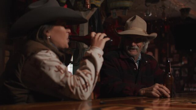Man and Woman Wearing Cowboy Hats Sitting at Bar Drinking Beer and Talking Inside of Old Rustic Saloon