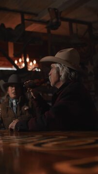 Man and Woman Wearing Cowboy Hats Sitting at Bar Drinking and Talking Inside of Old Rustic Saloon
