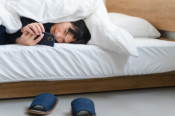 布団にくるまりスマートフォンを操作している若い男性　ライフスタイルイメージ