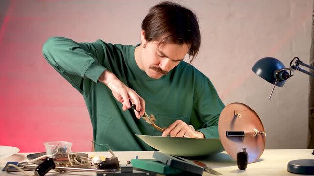 Concentrated man crafter repairing old electric lamp at home workshop at table, holding lamp base using pliers tool and trying to take off old fixed parts. Restoration hobby and upcycling.