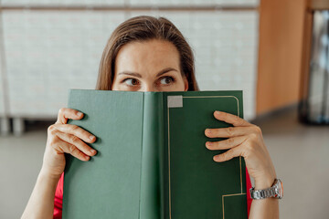 A young Hispanic woman with long brown hair holds a green folder in front of her face with humor.. She is in a modern coworking office environment, focused on her work.