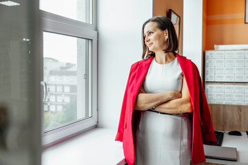 young Hispanic woman with short dark hair stands by a window in an office with crossed hands. She...