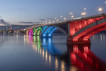The symbol of the city of Krasnoyarsk is an arched communal bridge over the Yenisei River in a multicolored illumination at night. Beautiful cities of Russia in the center of Siberia. High quality