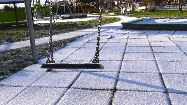 Children playground empty swing swining in slow motion back and fourth while covered in fresh white and fluffy first snow during a sunny day, green grass visible between the ice crystals and snow.