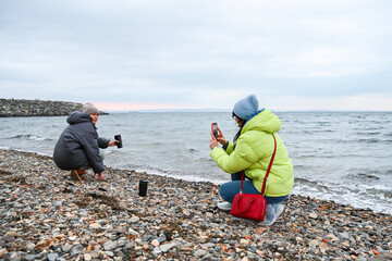 Two Russian women 50 and 60 years old by the sea in Vladivostok as one takes a phone photo of her friend enjoying calm mindful spring time outdoors