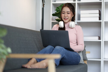 Comfortable young Asian woman wearing headphones around neck and working remotely from home, using laptop while sitting on sofa with legs up, enjoying relaxed home office environment.