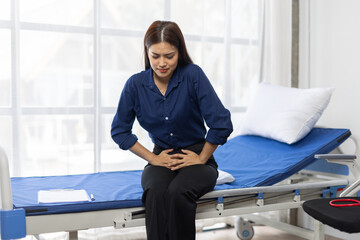 Young Asian woman sitting on hospital bed, experiencing severe abdominal discomfort, possibly due...